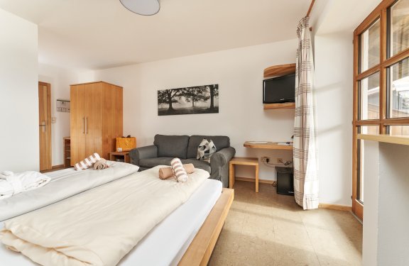 Helles Hotelzimmer mit zwei Betten, grauem Sofa, Holzschrank und Fenster mit Vorhang rechts