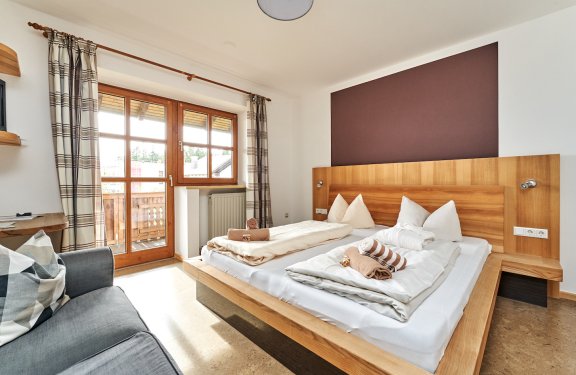 Helles Schlafzimmer mit Doppelbett, Holzkopfteil, Fenster mit Vorhängen und grauem Sofa links